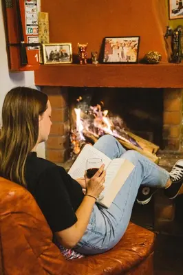 a woman sitting on a couch reading a book in front of a fireplace at Backpacker La Bo'M in Cusco